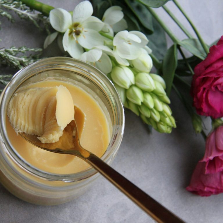 Pot de ghee maison doré posé sur une table fleurie, avec une cuillère un fond naturel