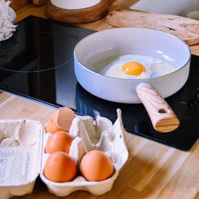 Œuf en train de cuire dans une poêle sur une plaque à induction avec une boîte d'œufs — exemple que Manger équilibré sans se ruiner est possible